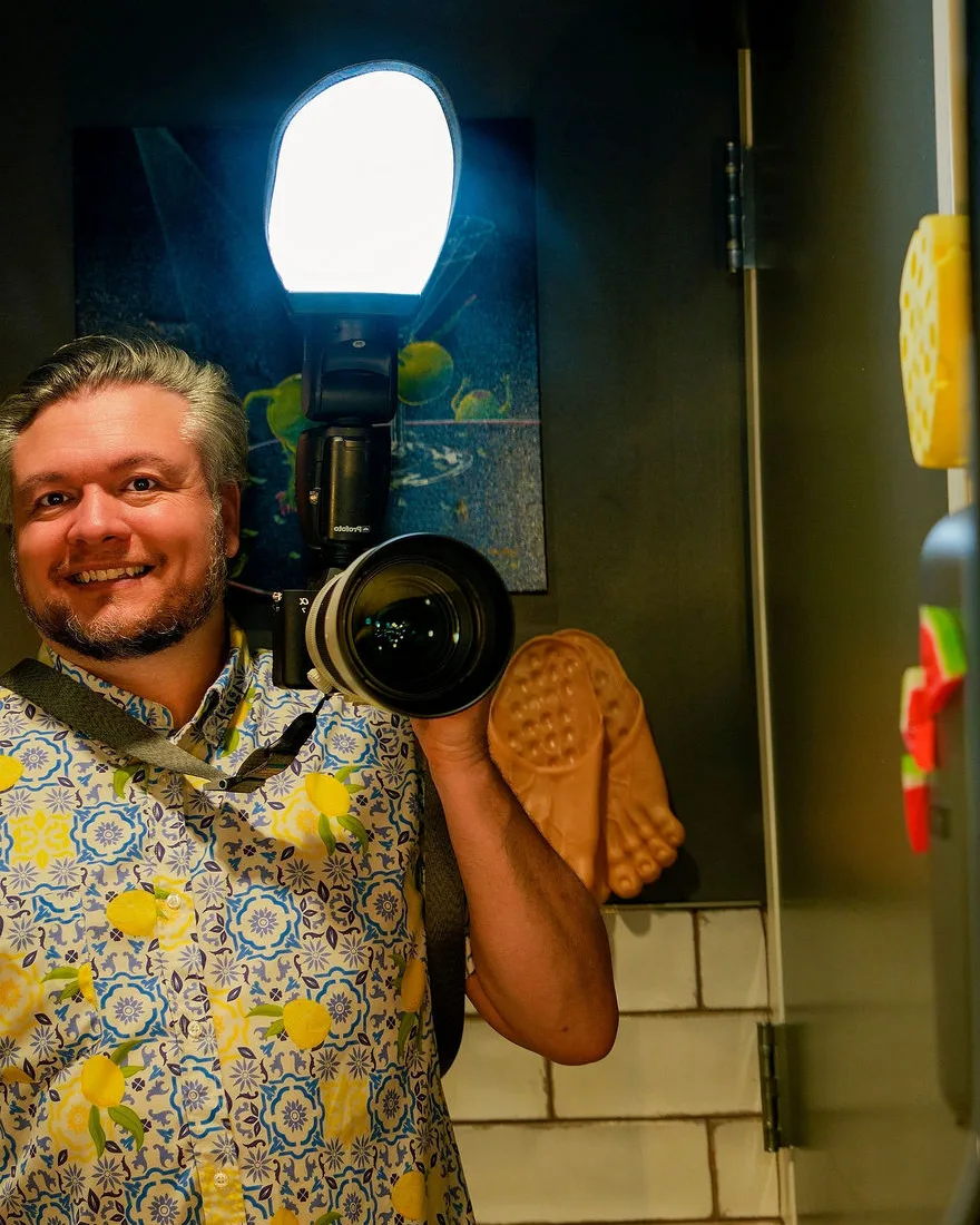 Greg Mulvey smiles into a mirror, holding up a camera with a long lens and a diffused flash.