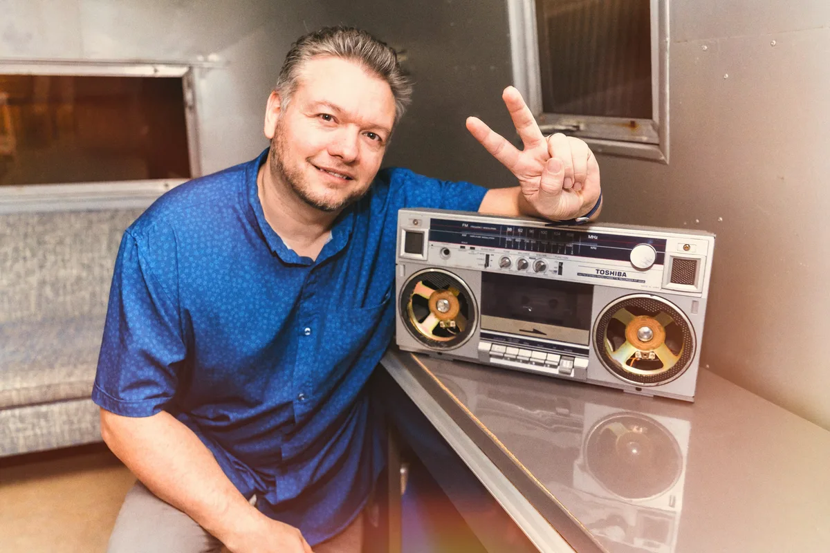 Greg Mulvey leaning on a counter beside a vintage boombox, one hand up in a peace sign.