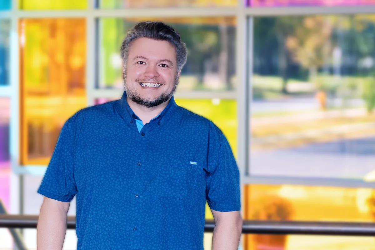 Greg Mulvey smiles in a blue patterned shirt in front of a wall of colored glass panels.