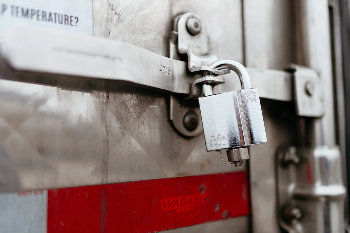 A chrome padlock locked through the hasp on a trailer's rear door, a red reflective strip below.