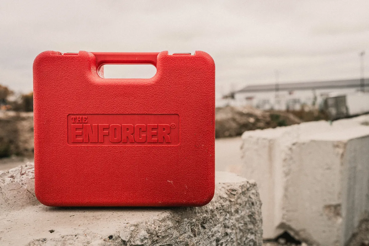 A red ENFORCER lock case standing on a concrete block, a trucking yard blurred behind it.