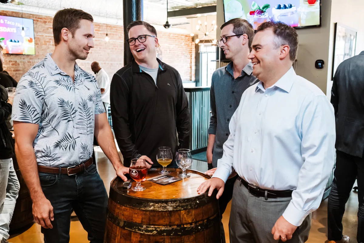 Four men in casual business wear talk and laugh around a barrel table in a brick-walled taproom.
