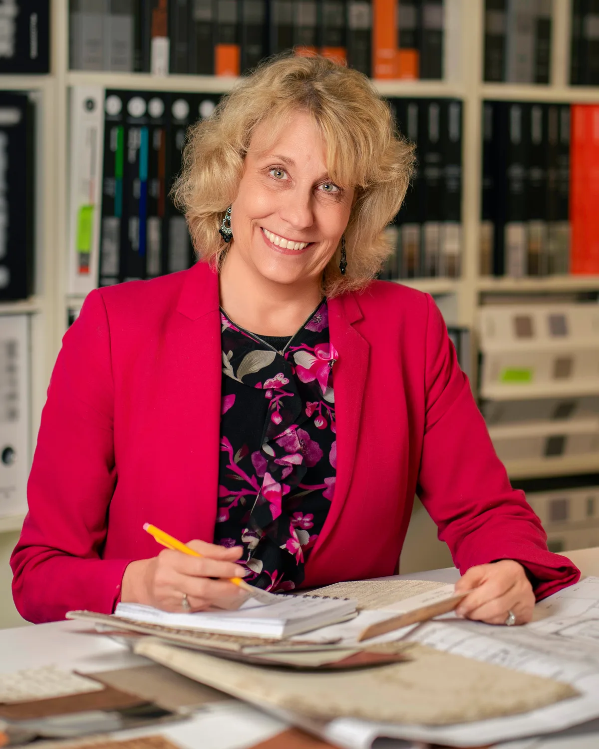 A designer in a red blazer smiles from a desk covered in fabric samples and drawings.