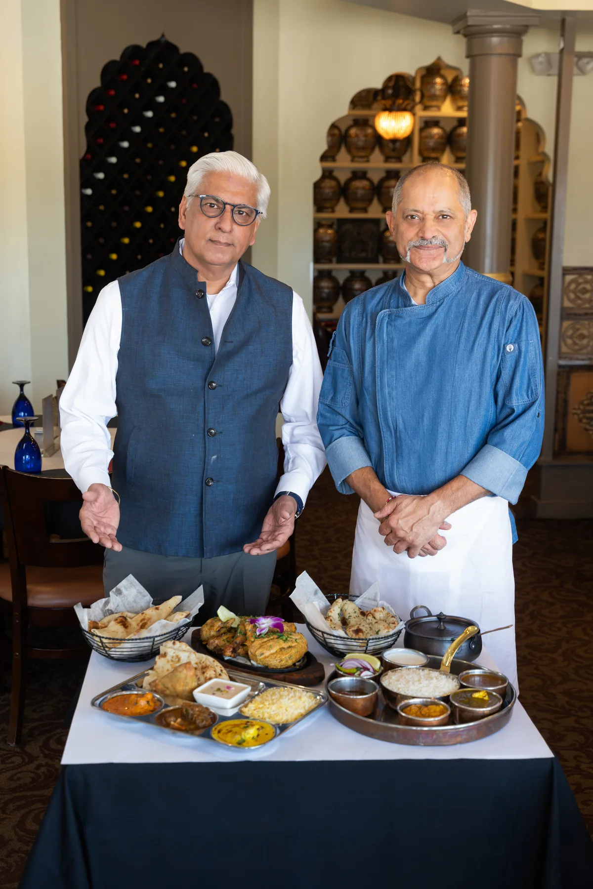 Two men, one in a chef's coat, stand behind a table spread with Indian dishes in their restaurant.