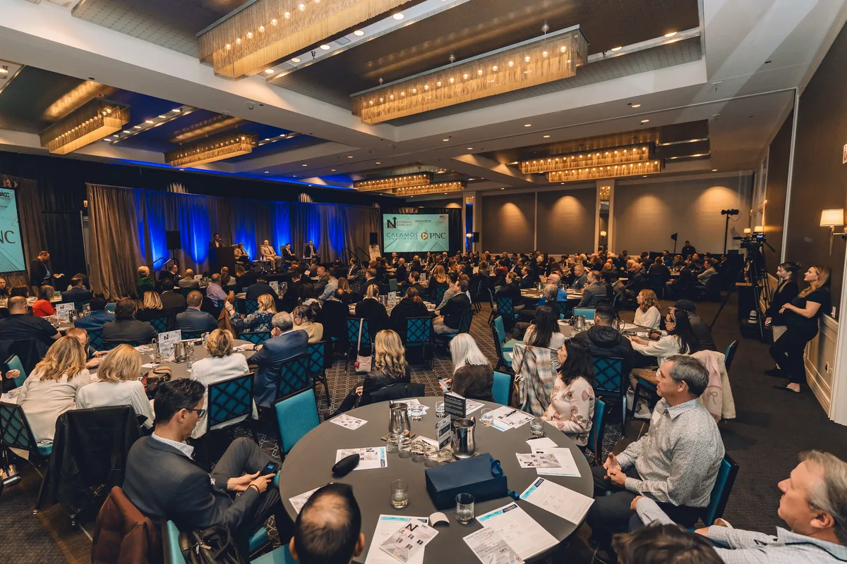 A full hotel ballroom of guests at round tables during a business luncheon, a presenter on the stage ahead.