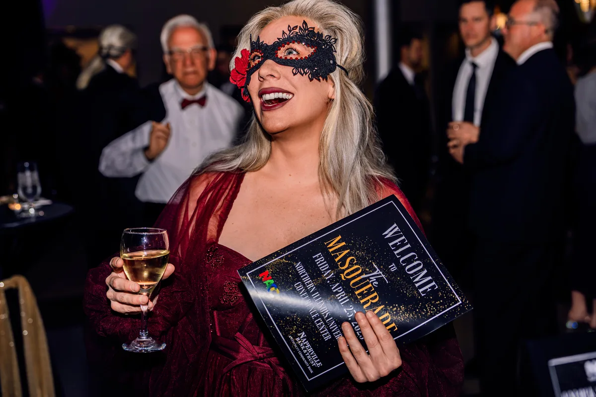 A guest in a lace mask and red dress holds a glass of wine and the program at a masquerade gala.