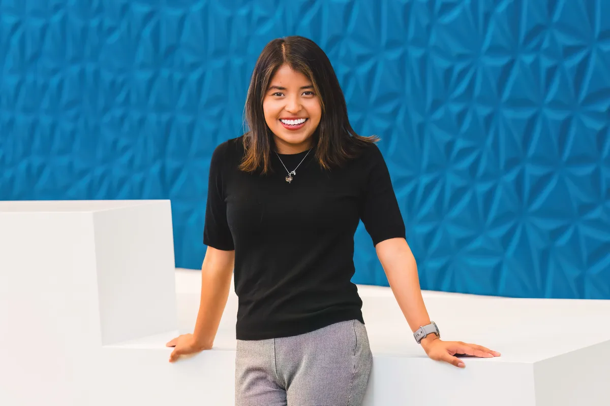 A woman in a black top leans on a white ledge against a sculpted blue wall for a location headshot.