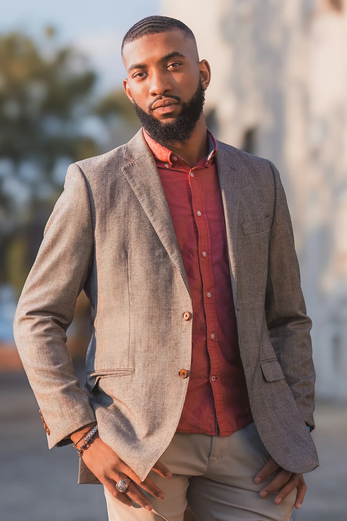 A man in a grey blazer and rust shirt, shoulders angled, photographed outdoors with a soft city background.