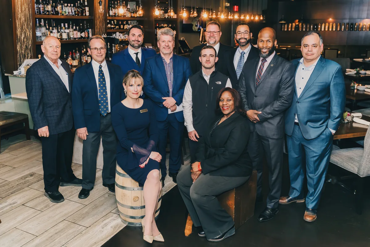 A group of business leaders gathered for a portrait in front of a lit restaurant bar.