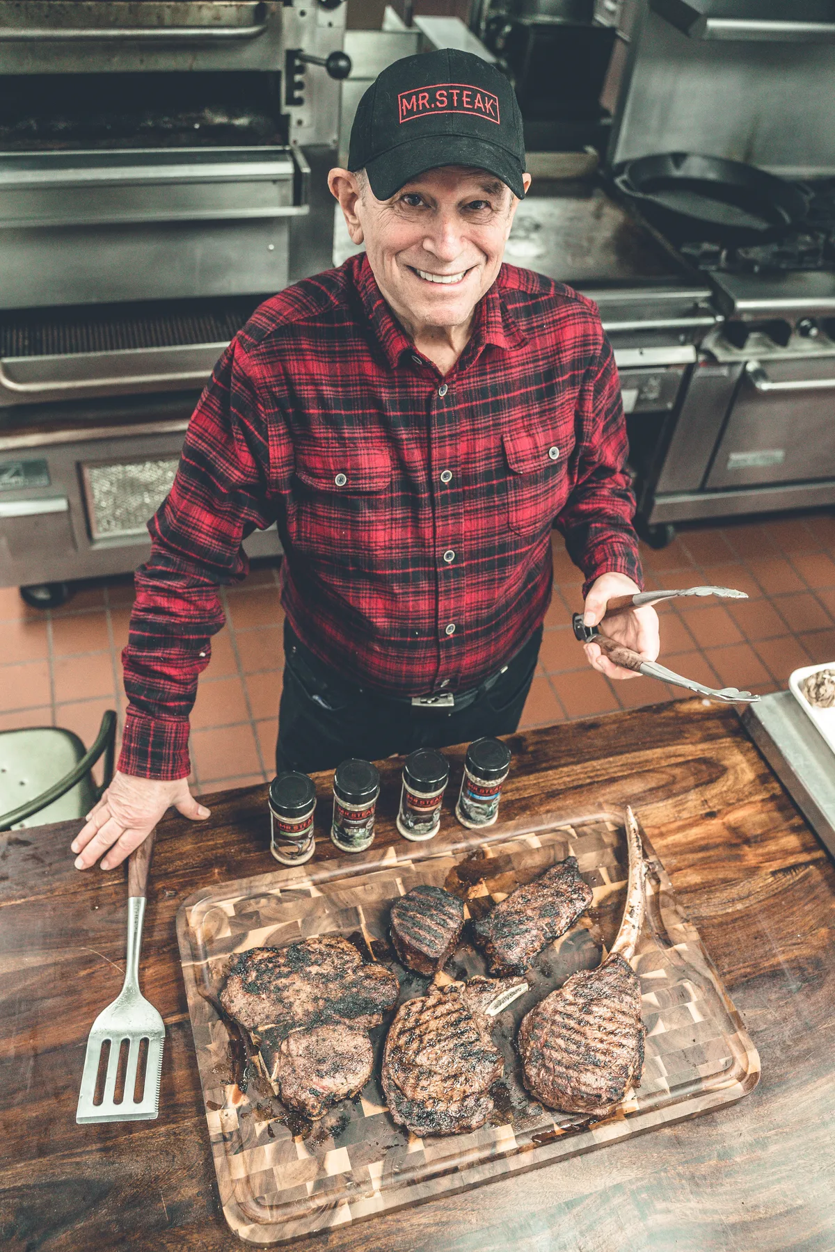 A man in a Mr. Steak cap and plaid shirt above a board of grilled steaks and seasoning jars.