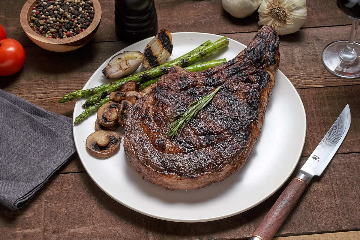 A charred bone-in ribeye with rosemary on a white plate, beside grilled asparagus, onion and mushrooms.