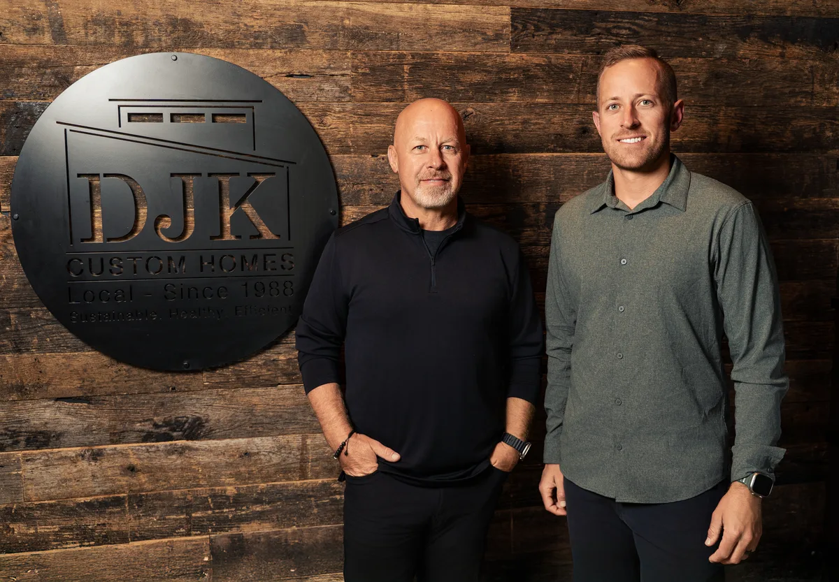 Two home builders stand in front of their company sign on a reclaimed wood wall.