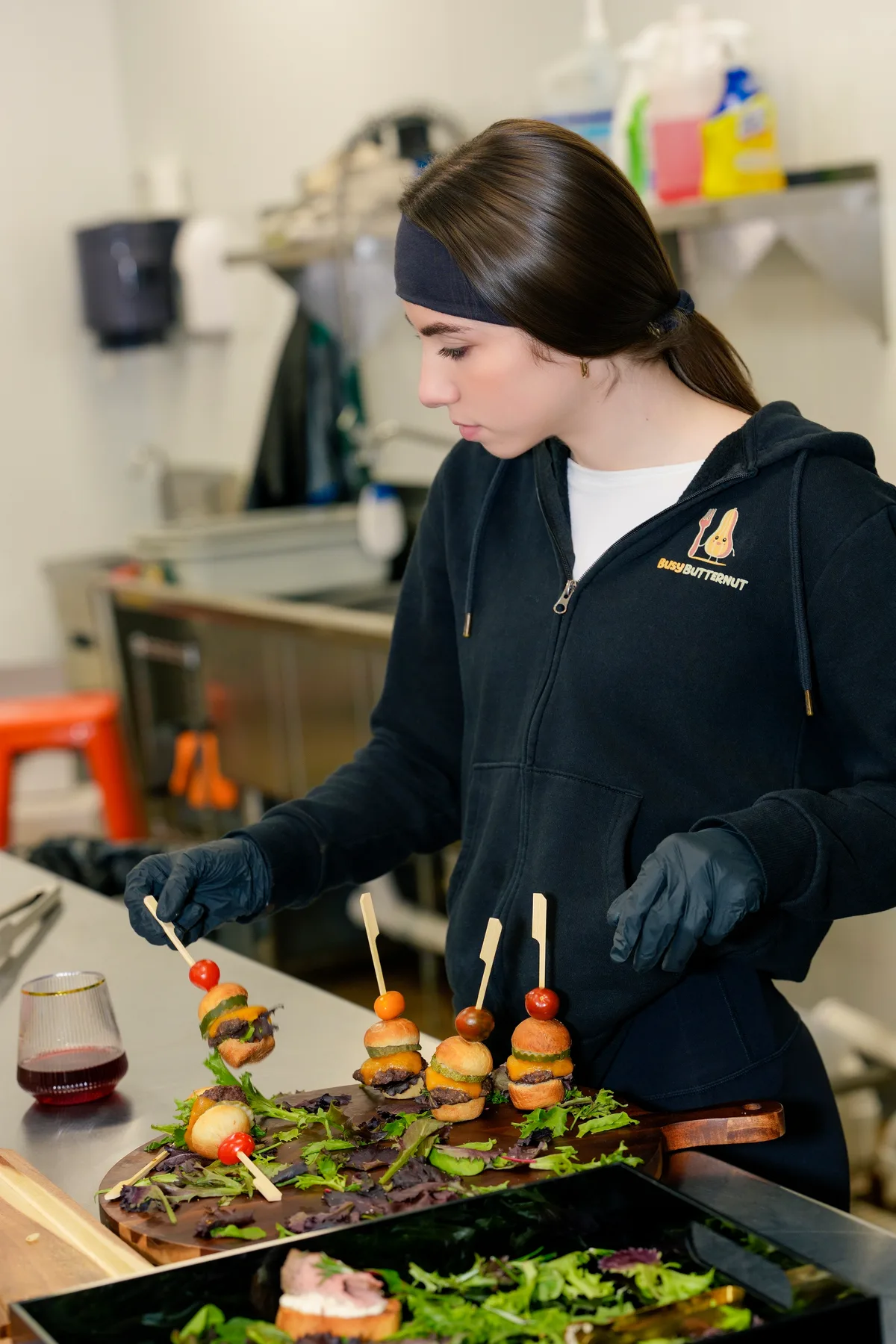 A cook in gloves plating skewers on a board at the bench of a commercial kitchen.