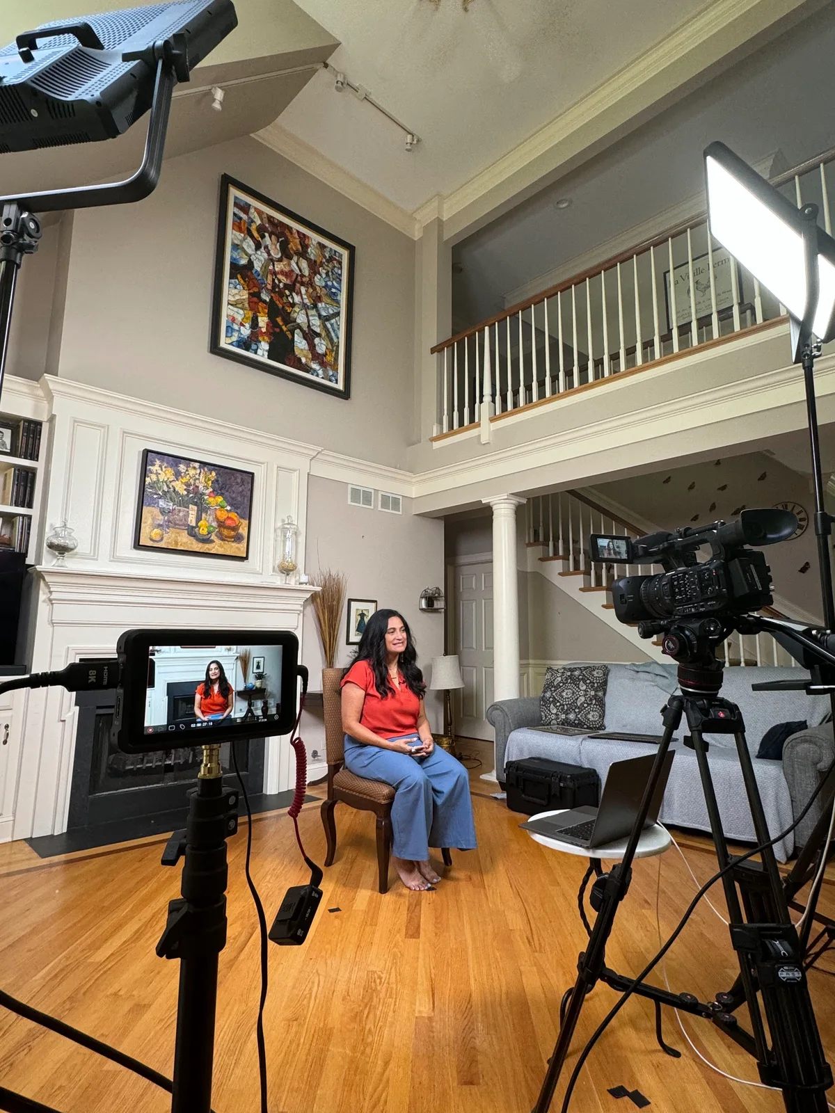 A woman sits for an on camera interview in a living room, the camera, monitor and lights set up around her.
