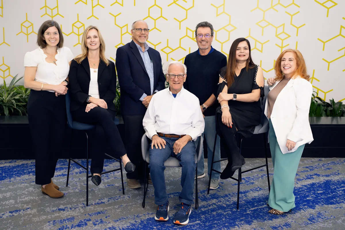 A seven-person team poses together in front of a bold yellow geometric wall.