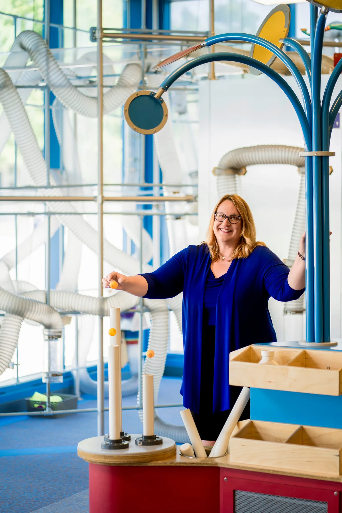 A museum leader smiles beside a colorful hands-on exhibit, holding up a ball to drop in.