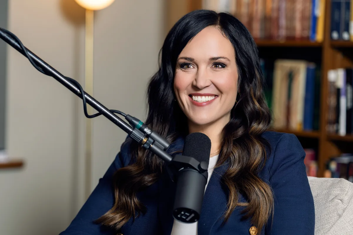 A podcast host at the microphone mid sentence, the studio bookshelves soft in the background.