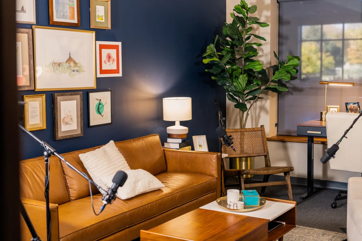 A podcast studio dressed as a living room: leather sofa, lamp, wicker chair and fig tree, microphones on boom stands.