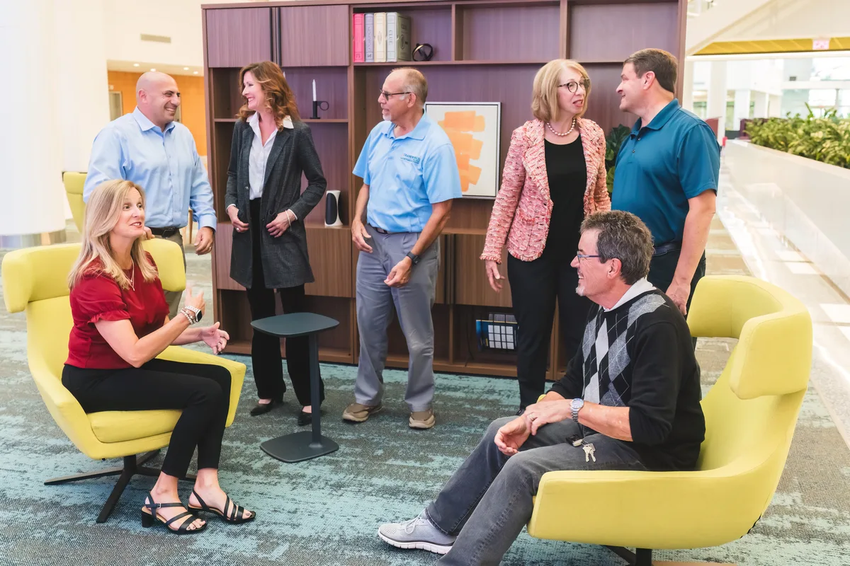 A business team meeting in an office lounge, some seated in armchairs and others standing in conversation.