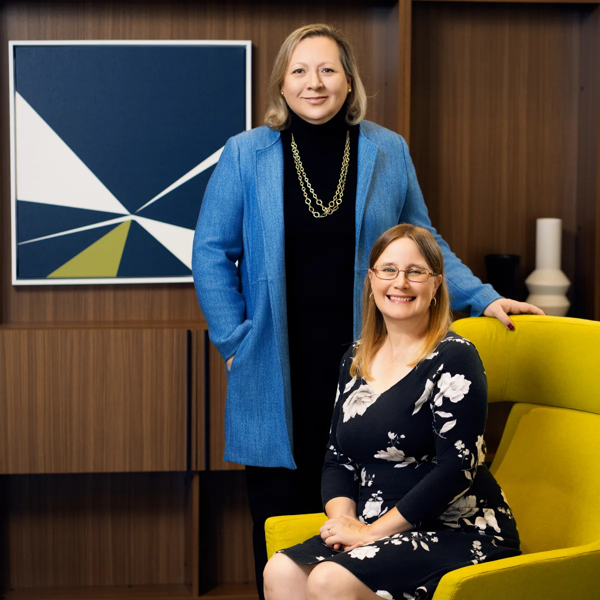 Two colleagues in a wood panelled office, one standing and one seated in a yellow armchair.