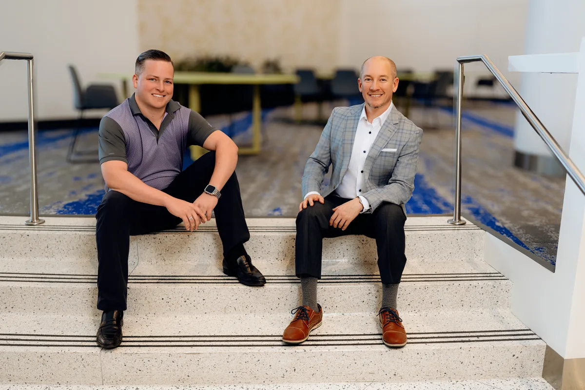 Two business partners sit on terrazzo stairs in a bright office lobby, smiling at the camera.