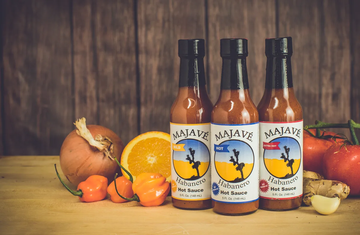 Three bottles of habanero hot sauce, mild, hot and extra hot, on a wooden table with habaneros, an orange and tomatoes.