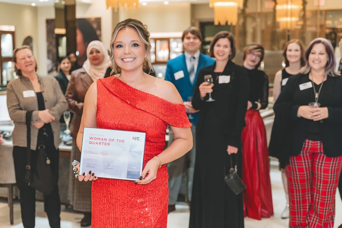 An award recipient in a red dress holds up a certificate, surrounded by applauding guests.