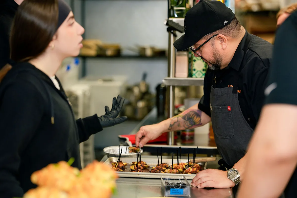 A chef in a black cap and apron placing bacon-wrapped appetizers on a sheet tray as a guest talks beside him.