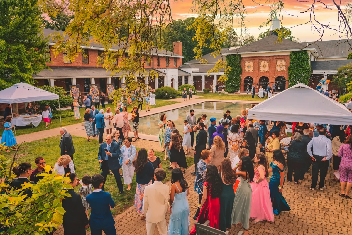 Guests in formalwear filling a brick courtyard around a reflecting pool at sunset, white tents and string lights overhead.