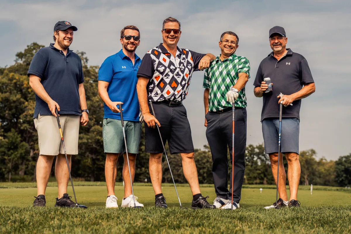 Five golfers in polos lined up on a tee box, grinning, clubs planted on the grass.