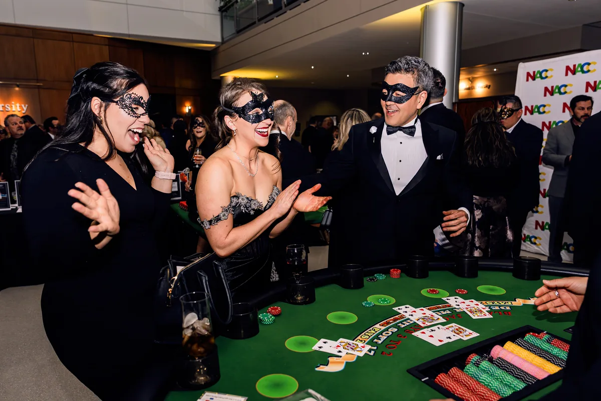 Guests in masquerade masks and evening wear cheering around a blackjack table at a gala.