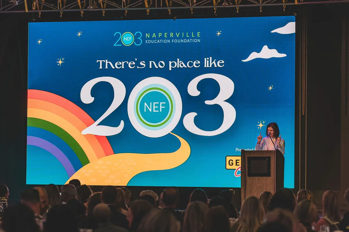 A speaker at a podium beside a giant screen reading There's no place like 203, the audience in silhouette.