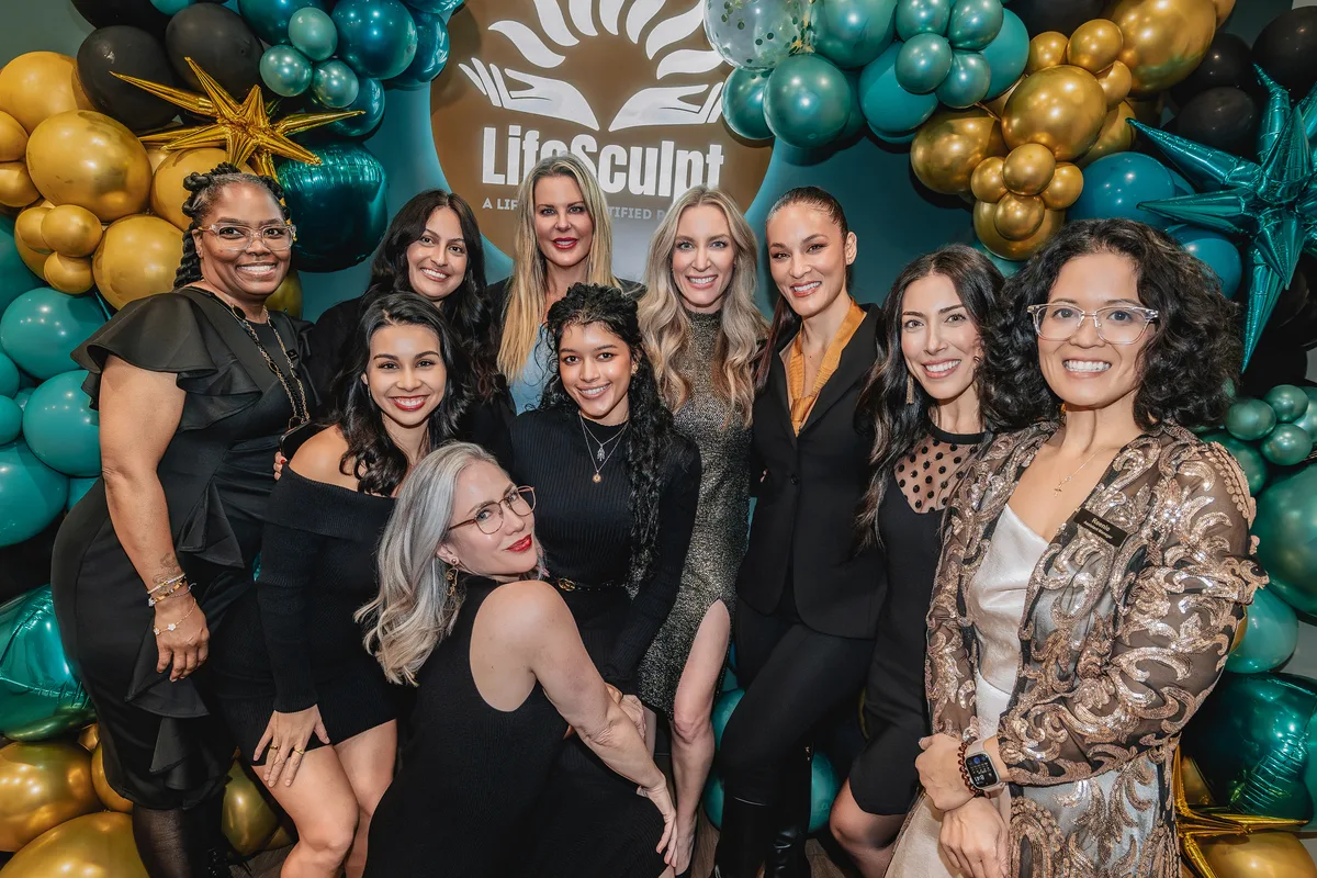 Ten women in evening wear smiling together in front of a teal and gold balloon arch and a LifeSculpt sign.