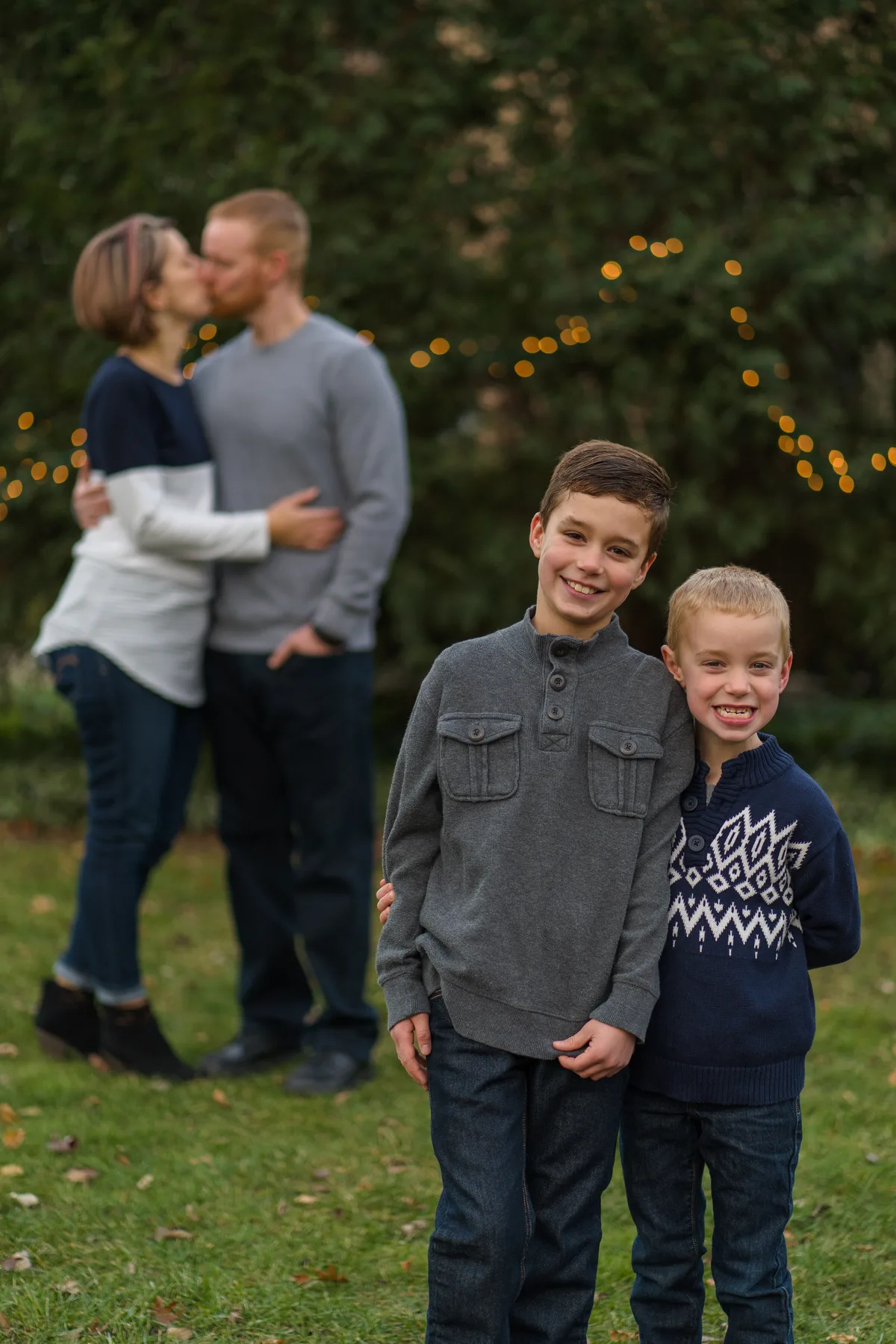 Two brothers grin at the camera while their parents share a kiss behind them, string lights glowing in the hedge.