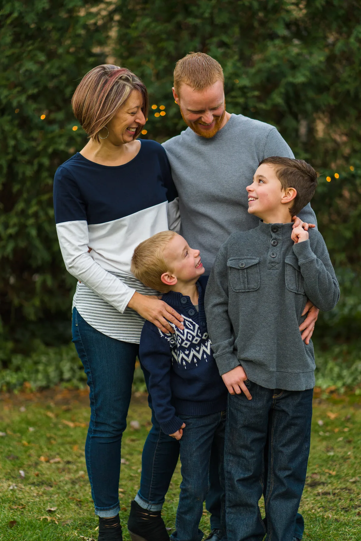 Two brothers look up at each other and laugh while their parents smile down at them in front of a dark green hedge.