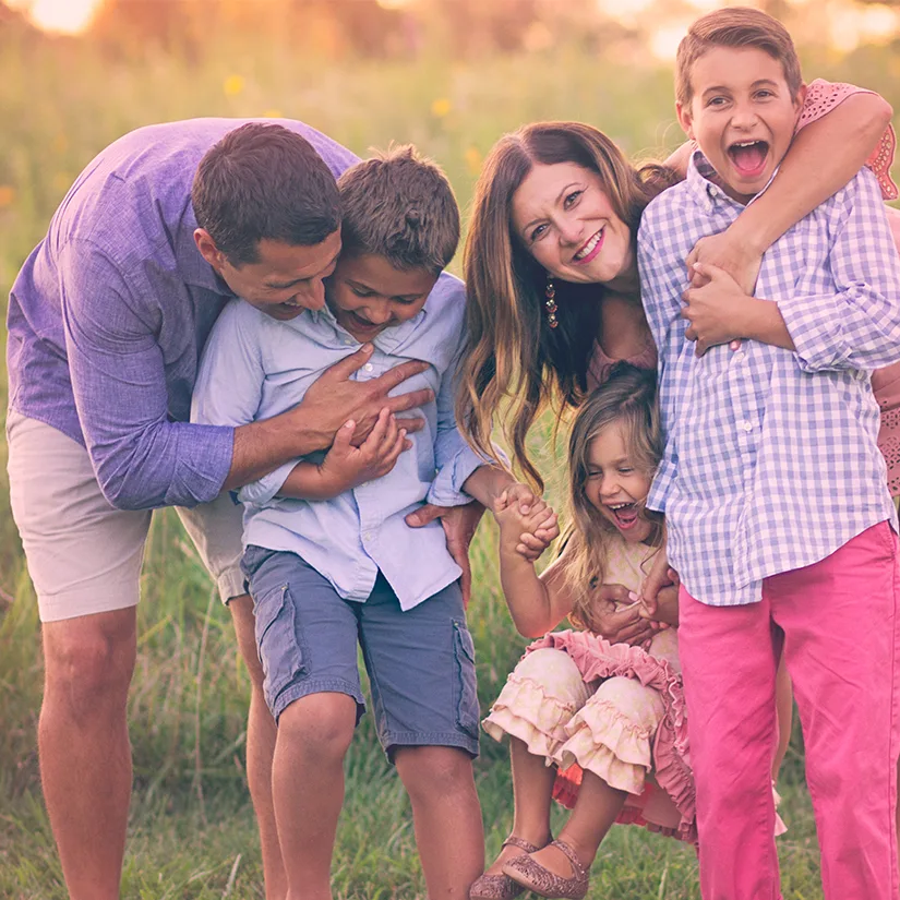 A family of five laughs and piles together in tall grass in warm evening light.