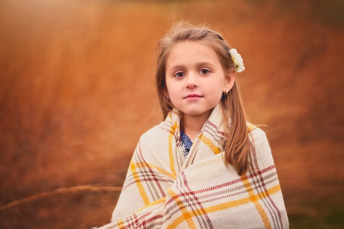 A young girl wrapped in a plaid blanket looks straight at the camera, tall autumn grass glowing behind her.