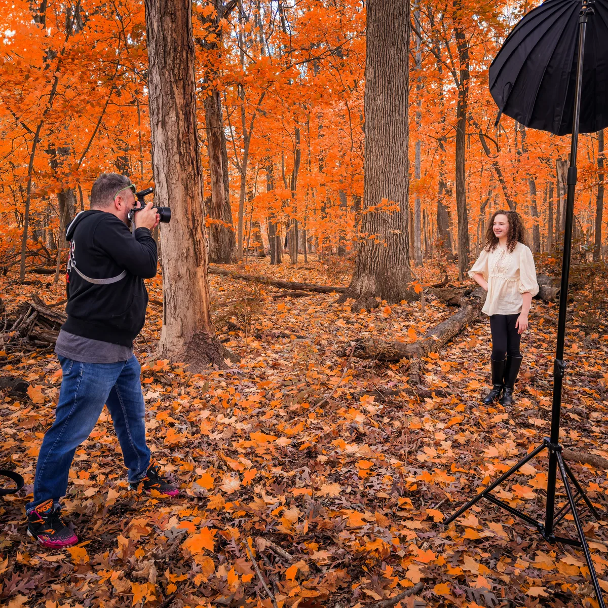 Greg Mulvey photographs a smiling teenager in autumn woods, a softbox on a stand beside her.