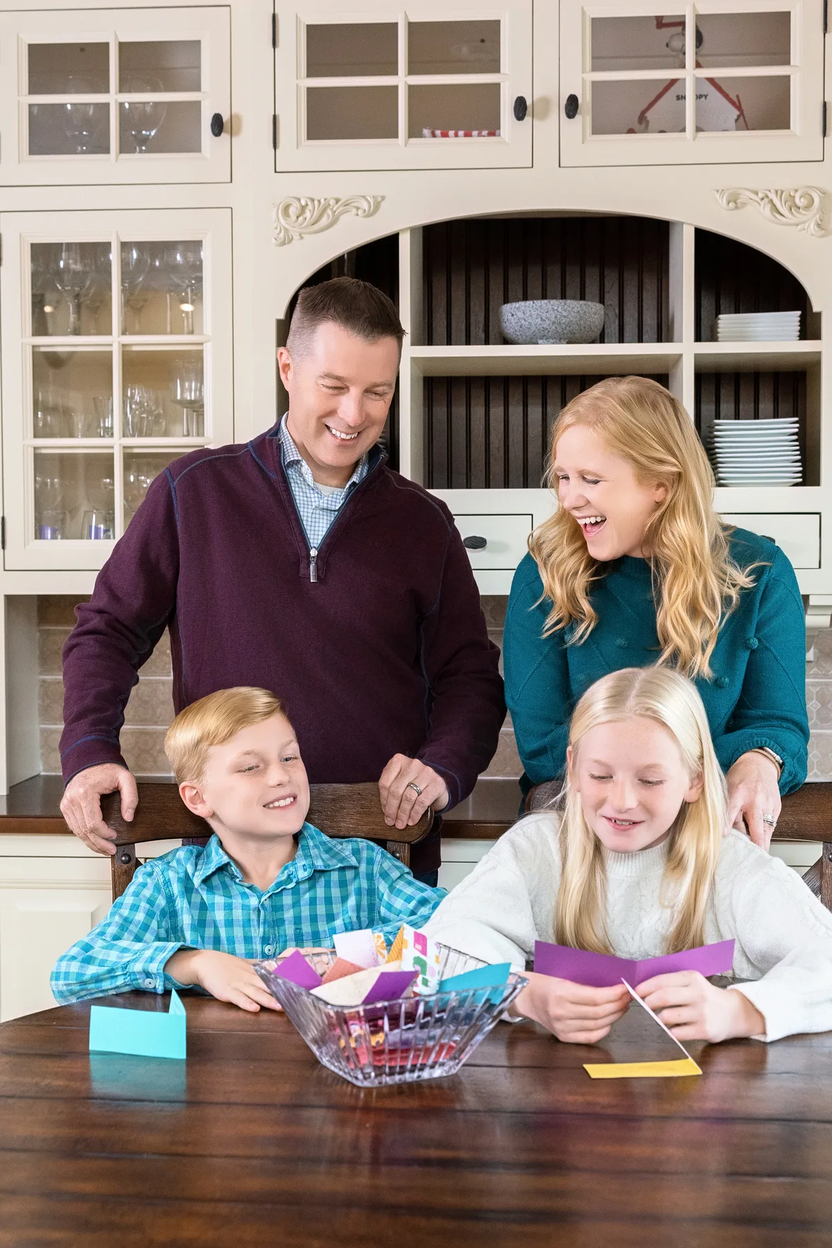 Two parents lean over their kids at the kitchen table, everyone laughing over a bowl of folded notes.