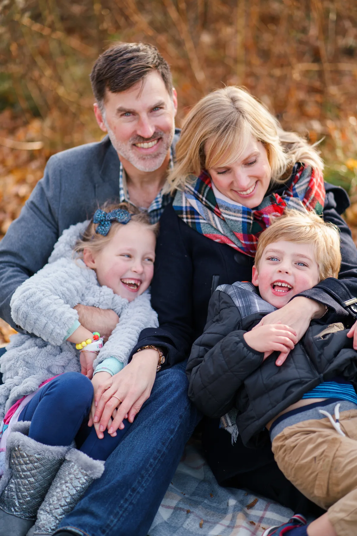 A family of four tumbles together on a blanket in autumn leaves, both kids laughing in their parents' arms.