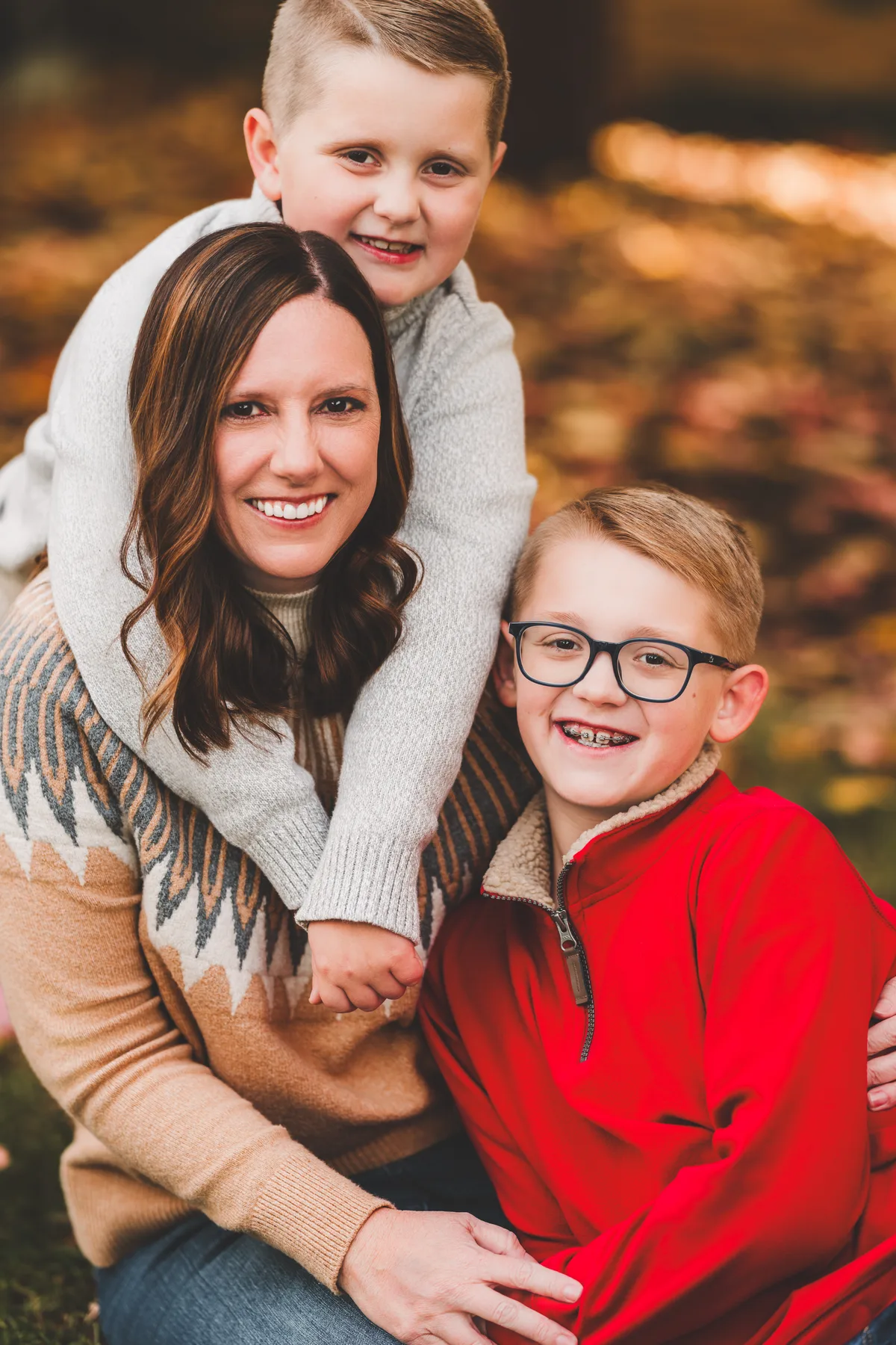 A mother sits in autumn leaves with one son draped over her shoulders and the other leaning in.