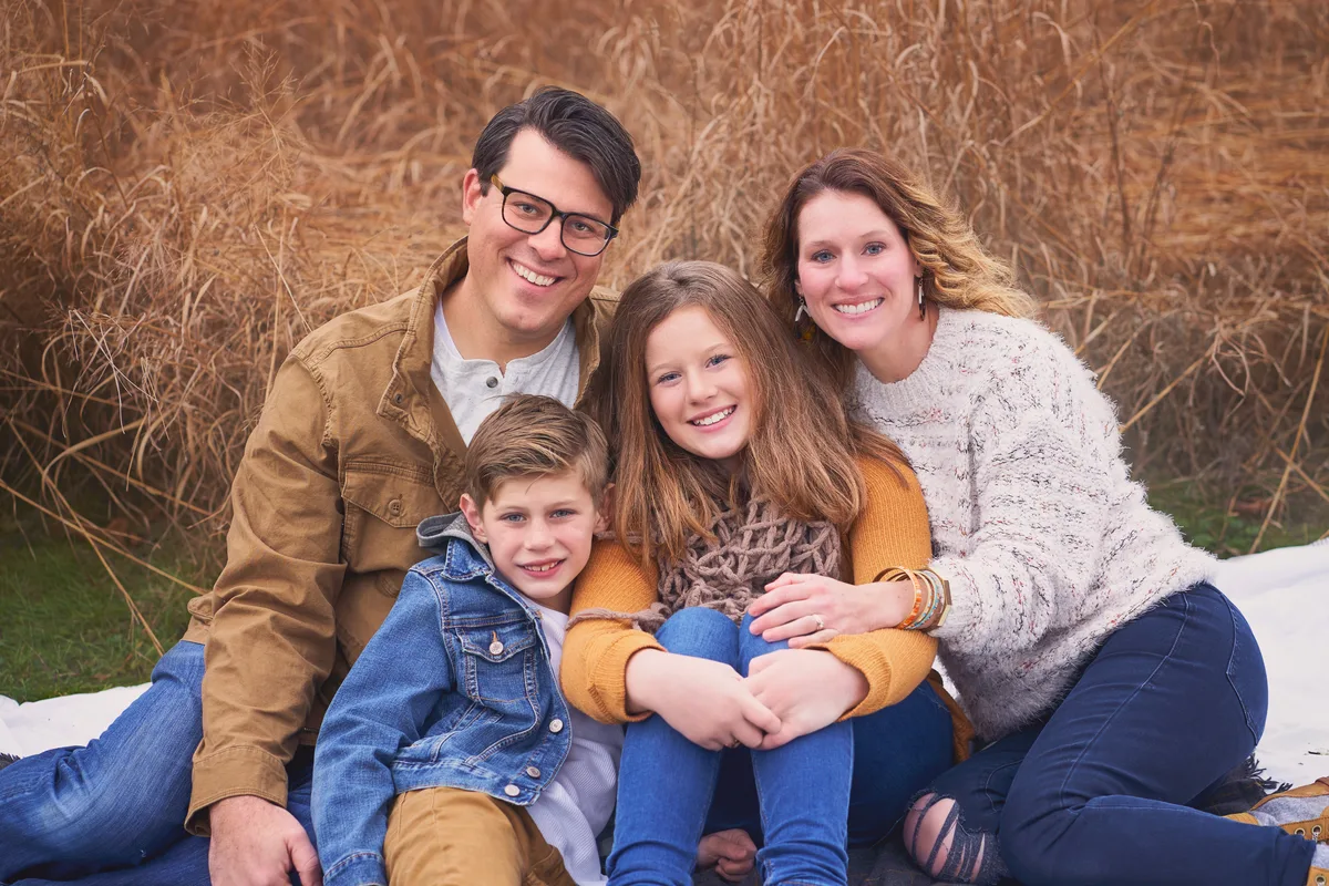 A family of four sits close together on a blanket in front of tall autumn grass.