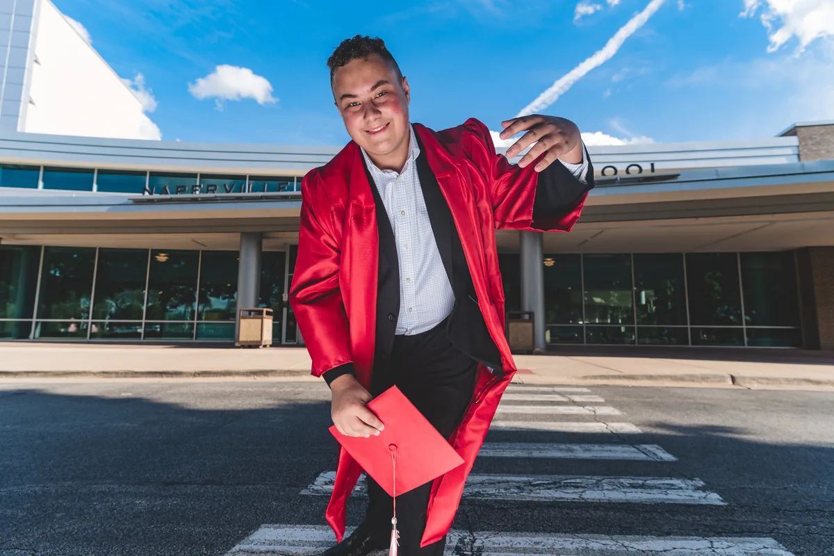 A high school senior in a red graduation gown leans toward the camera on a crosswalk, cap in hand.