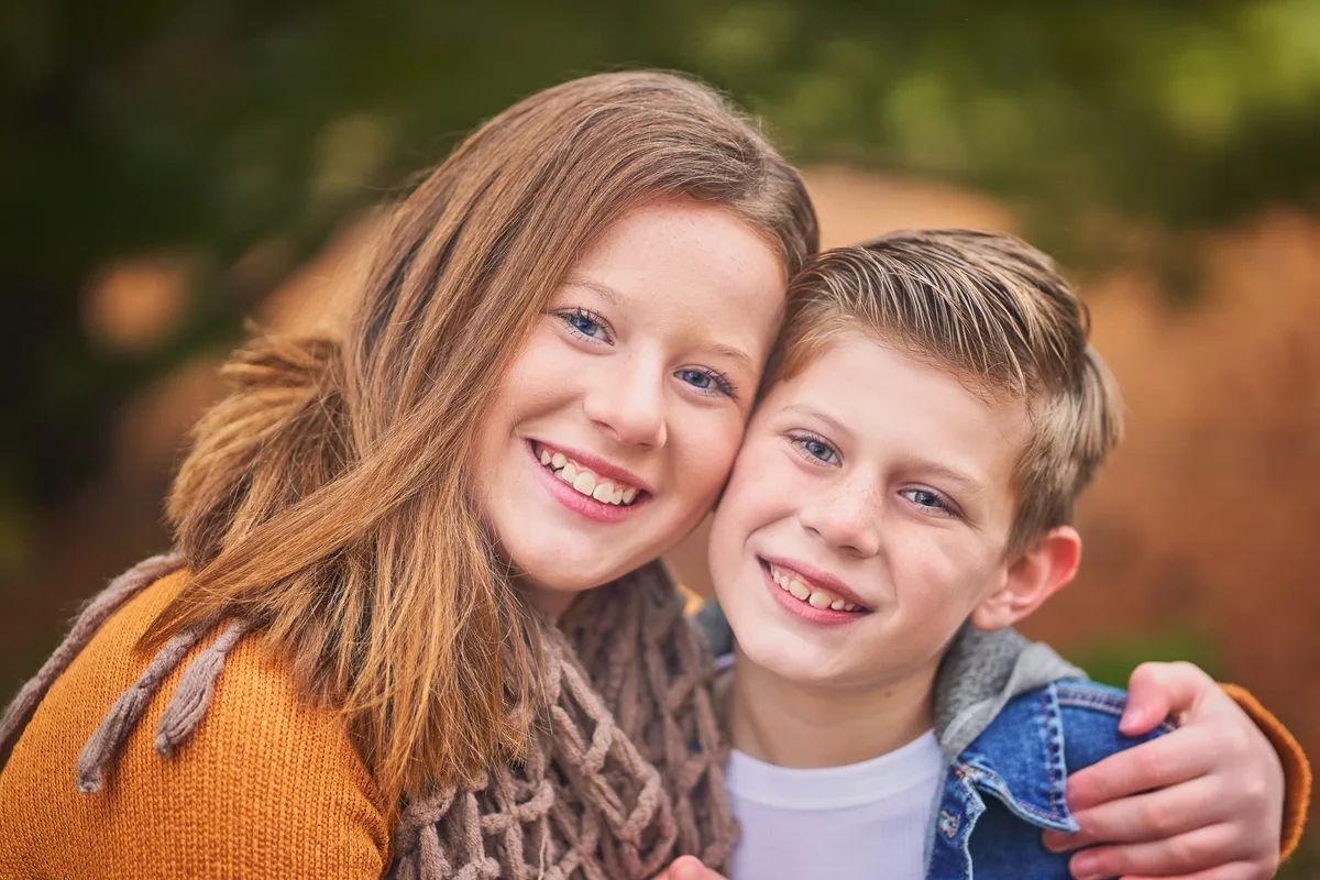 A sister and brother press cheeks together and smile, autumn color soft behind them.
