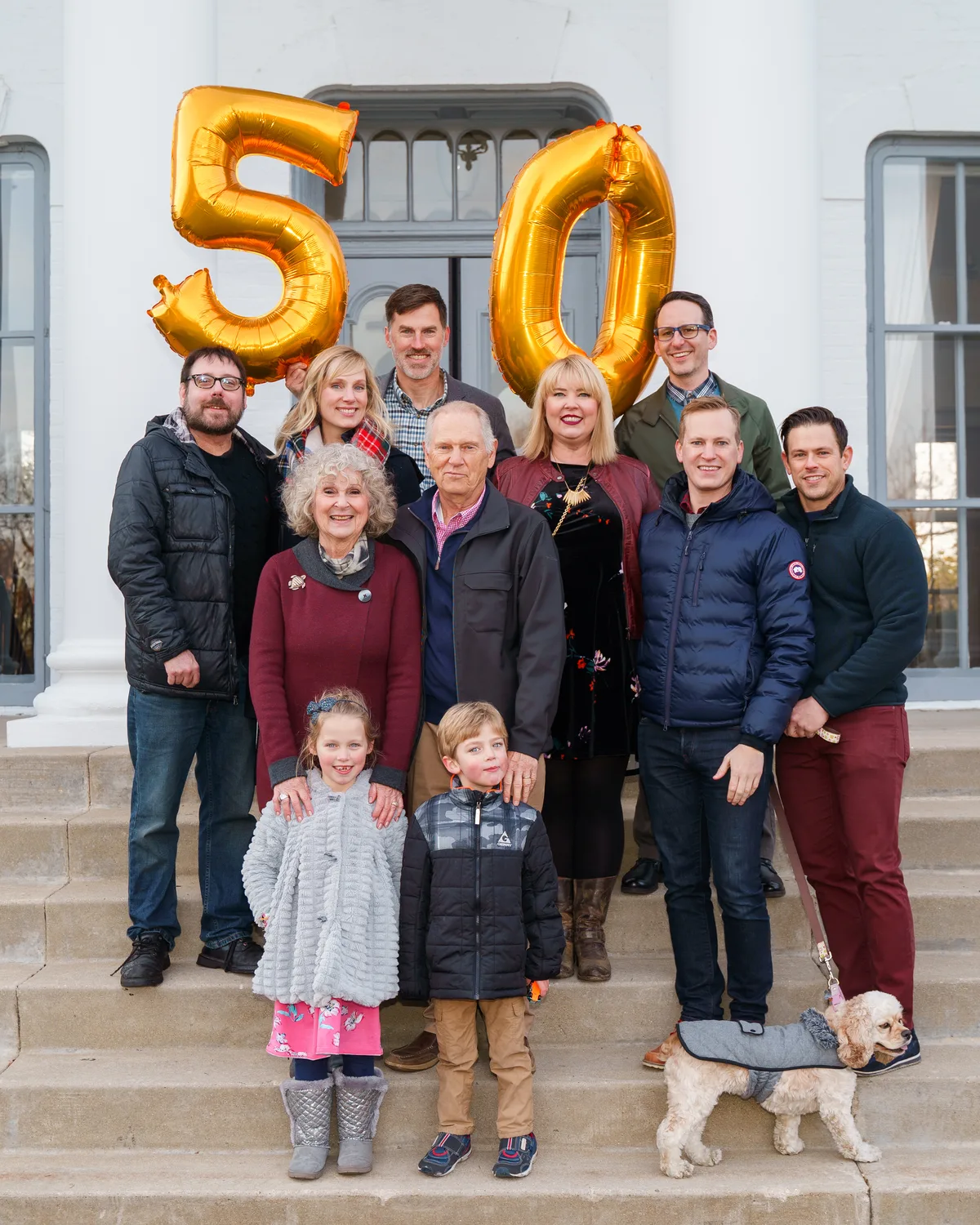 Three generations gather on the steps of a white columned building under gold 50 balloons, the family dog at their feet.