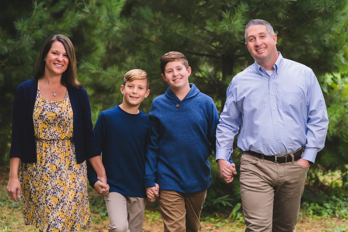 A family of four walks hand in hand in front of a stand of pine trees.