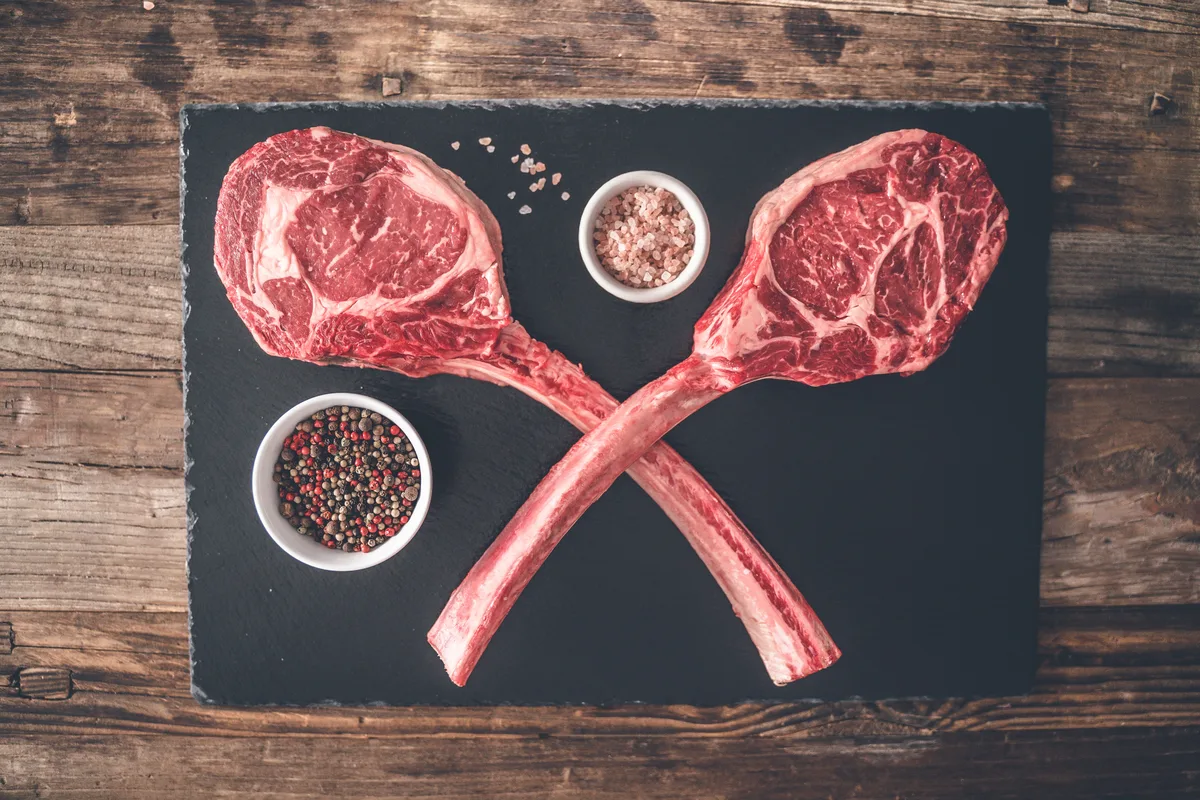 Two raw tomahawk ribeyes crossed on a slate board between small bowls of pink salt and peppercorns.