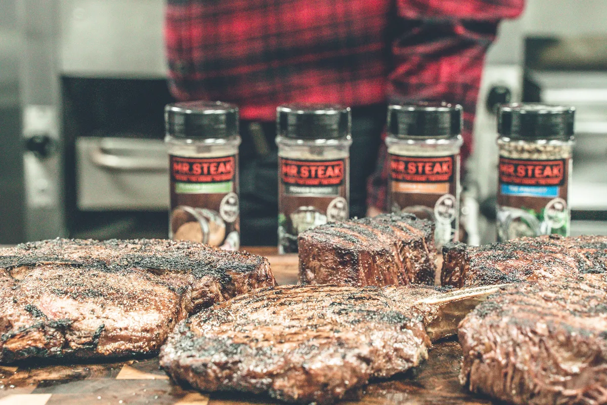 Grilled steaks on a cutting board in front of four Mr. Steak seasoning shakers.