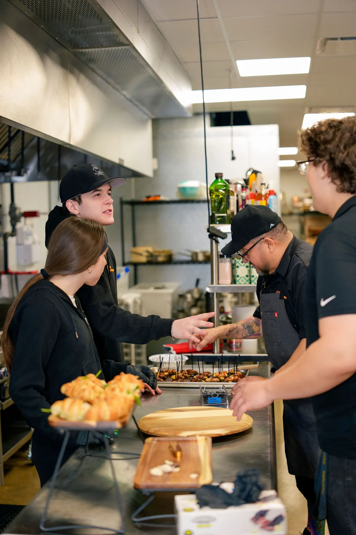 The Clarify Kitchen team talks across a steel prep counter, trays of skewered bites and croissant sandwiches ready.