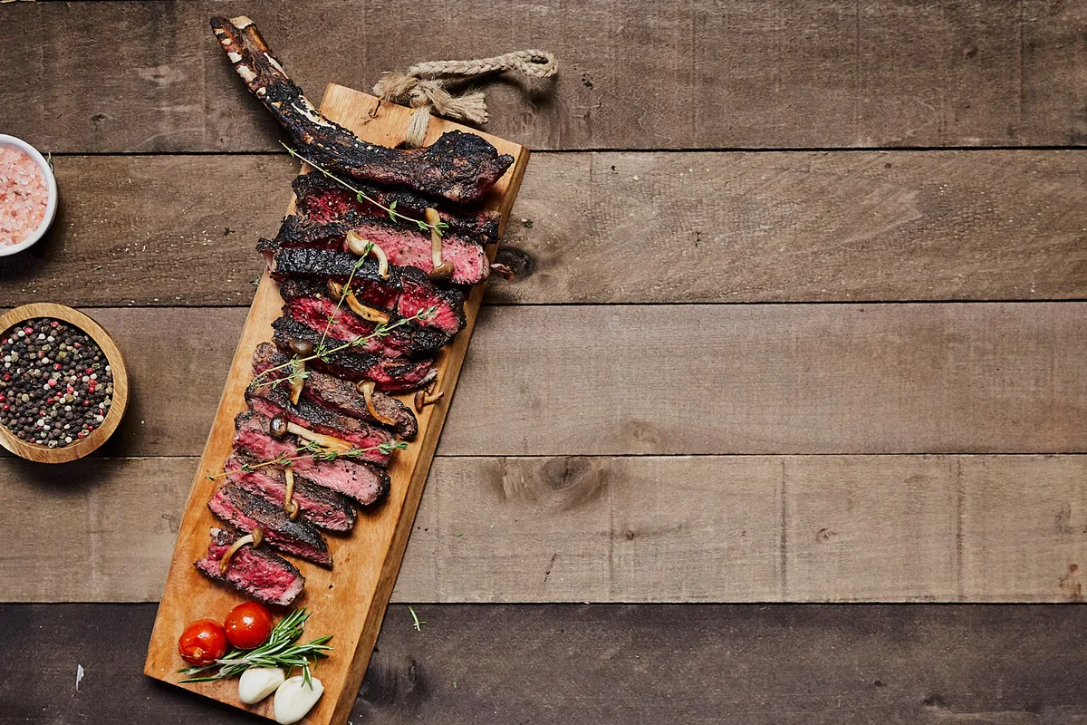 A sliced tomahawk steak fanned on a wooden board with mushrooms, thyme, cherry tomatoes and garlic, shot from above.